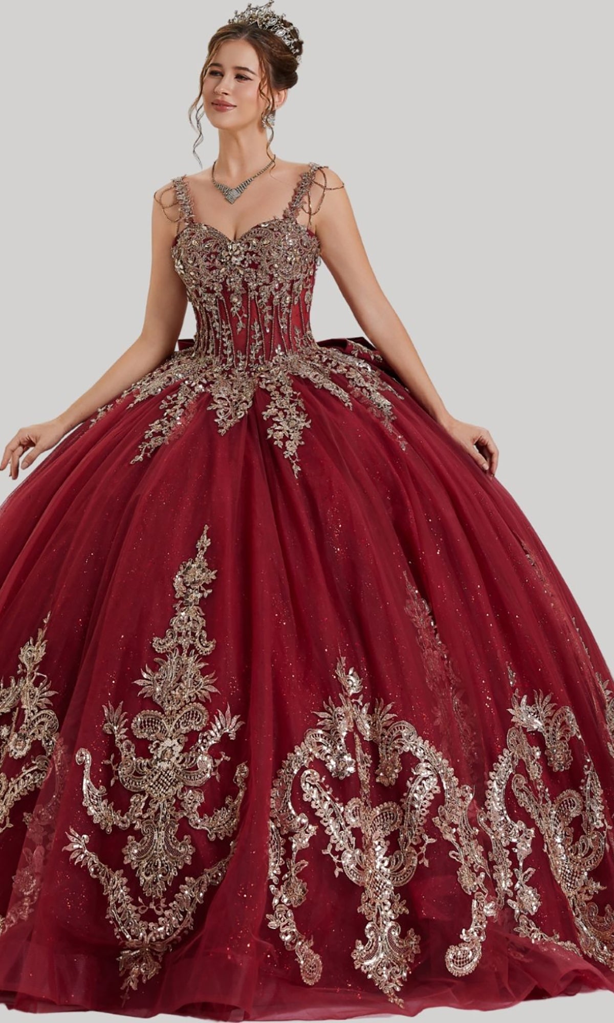 A woman wearing the Quinceanera Dress 26407 by Stella Couture in burgundy, featuring gold embroidered appliques, a glitter tulle skirt, and a tiara stands against a plain gray background.